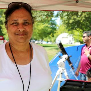 An Indian woman facing the camera and smiling. Her hair is tied back and she wears sunglasses on top of her head. She stands under a canopy-like tent, some of which can be seen running horizontally across the top of the photo. In the background is a sunny field with trees and a man standing beside a telescope.
