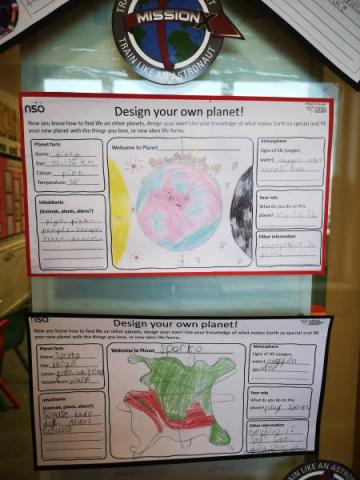 Two student-designed planets displayed on a wall, each with a worksheet titled "Design Your Own Planet." One planet is colored in pink and yellow, while the other is green and red