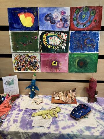 Part of an art exhibition. A table in the foreground has a tye-die cloth covering it and various handmade aliens and rockets. Behind the table are 9 paintings inspired by astronomical observations