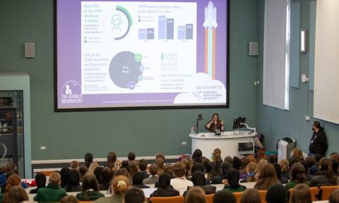 A presenter speaks to a large audience of students in a lecture theatre, with a slide about diversity in the STEM workforce displayed on a large screen behind them.