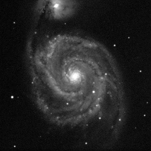 A large spiral galaxy, viewed face-on, with some white dots of stars visible in the background. The centre of the galaxy has a small, white sphere that glows brightly. Spiral arms in various shades of grey extend away and wrap around it. Slightly to the left of centre, at the top of the image, one of the spiral arms extends up towards a smaller galaxy. This also has a bright centre, and dust surrounds it in an oval cloud.