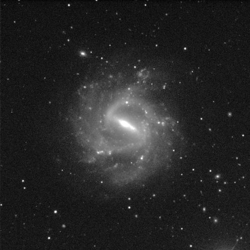 A spiral galaxy angled face-on against a black background with white dots of stars. The galaxy has a small, circular, bright, white centre and dusty grey spiral arms sweeping around it. It is surrounded in a grey, cloud-like fuzziness. This cloud forms an almost bar shape across the centre of the galaxy, though it is obscured by the centre shining brightly.