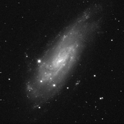 A spiral galaxy against a black background with white dots of stars scattered throughout. The galaxy has a small bright white centre and is surrounded by fuzzy grey dust, resembling a cloud. The shape is an elongated oval stretched towards the lower left and upper right of the image.