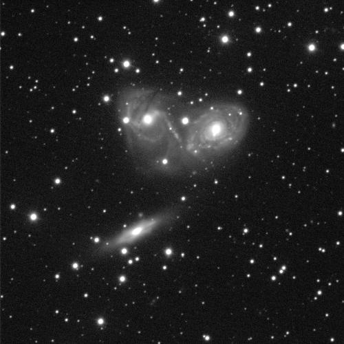 A black background with white dots of stars. There are 3 small galaxies centered in the middle of the image. Two are next to each other with one below these. The galaxy's have bright white, circular centres surrounded by grey cloudy dust. The top left has defined spiral arms that extend up and down while the top right has spiral arms wrapping around it entirely. The bottom galaxy is angled almost side-on, making the grey dust a thin disc shape.