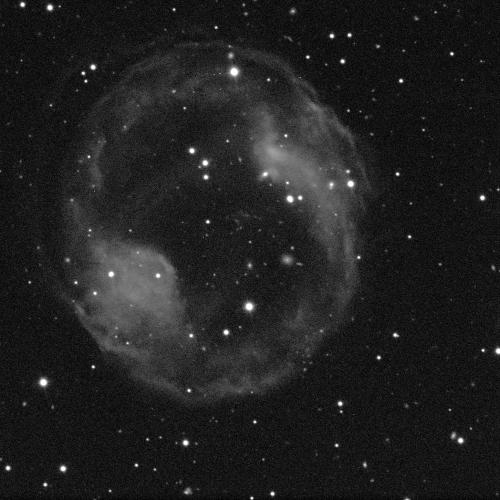 A ring-shaped nebula against a black background with some small white dots of stars. The nebula is cloud-like, grey, and fuzzy.