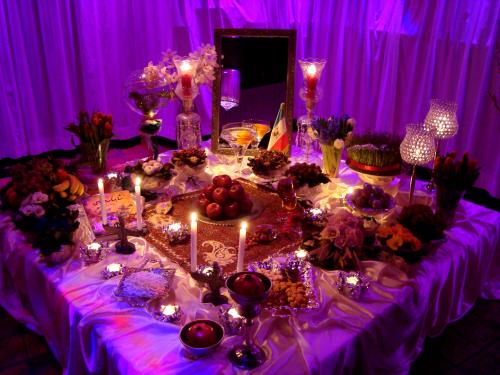 A clothed table illuminated by purple light. There are various dishes and food and decorations such as a rectangular mirror, lit candles in holders, and vases of flowers.