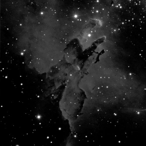 A grey, cloudy nebula against a black background with small dots of white stars. The nebula covers most of the image, but there's a dark patch of the night sky towards the lower left corner. In the middle of the image, pillar-like structures of dust rise up towards the top-right corner.