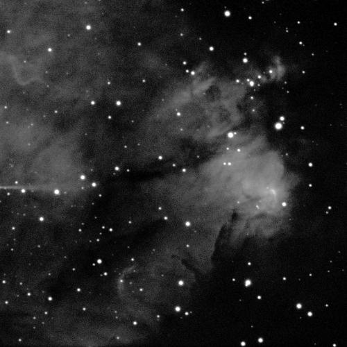 A cloudy nebula against a black background with white dots of stars scattered across the image. The nebula is various shades of grey, with the brightest part towards the middle-right of the image. To the right of this part, a dark patch of sky extends almost vertically. The nebula spreads towards towards the upper-left and lower-right, in a wide funnel-like shape.
