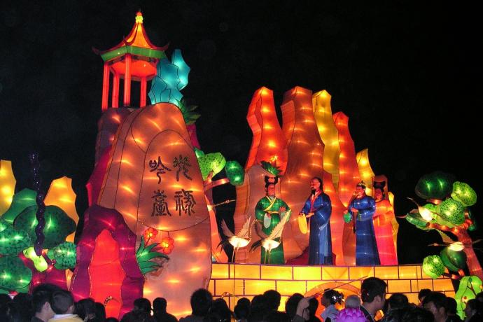 A photo of a stage showing a set made of illuminated pieces. It depicts a landscape, showing tall mountains with a structure on top of one and some trees. Three people stand on a lit up block wearing robes.