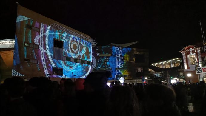 A photo of a crowd at night with buildings on the left. Patterns of light are being projected on to screens on the buildings.